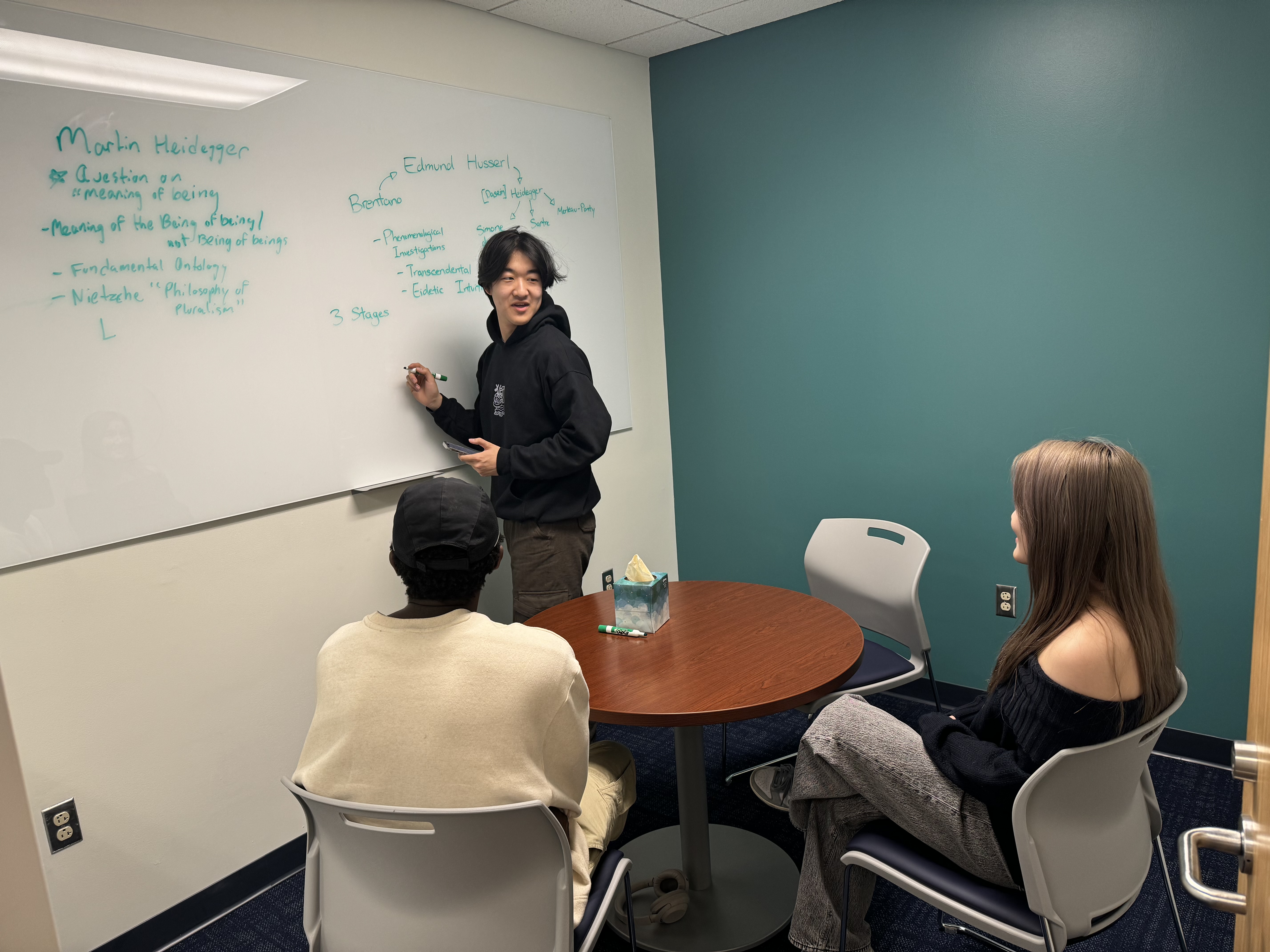 Three students studying together in a group study room. One student is writing on a white board