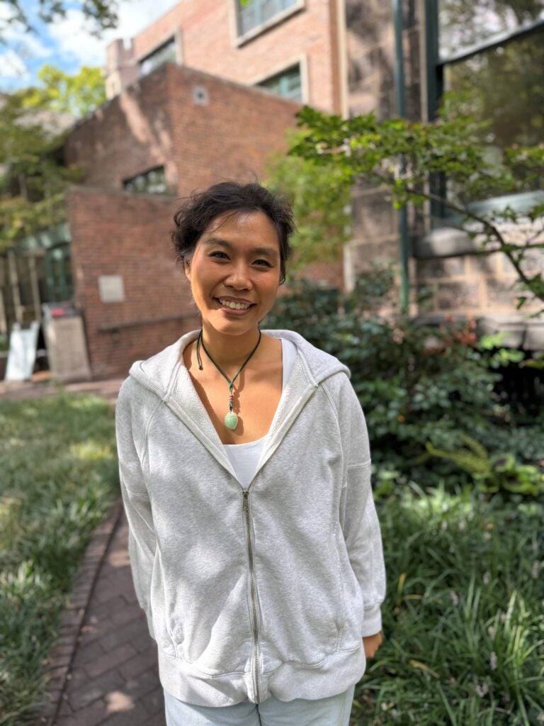 A young woman with dark hair and olive skin wears a white sweater and stone necklace. She stands in front of greenery and a brick building.
